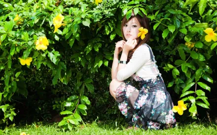 A woman named Mikako Zhang Kaijie poses among lush greenery and bright yellow flowers, creating a vibrant and charming HD desktop wallpaper background.