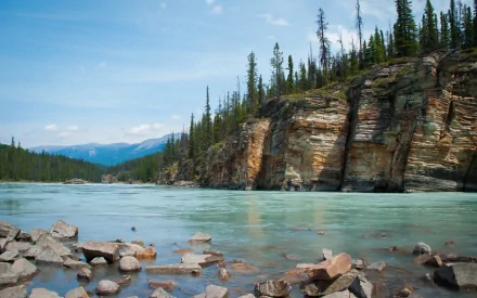 HD desktop wallpaper showing a serene nature scene with a rocky cliff beside a calm river surrounded by pine trees under a clear blue sky.