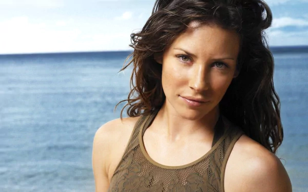 HD PC desktop wallpaper featuring celebrity Evangeline Lilly with wavy hair, wearing a sleeveless top, standing in front of a calm sea under a clear sky.