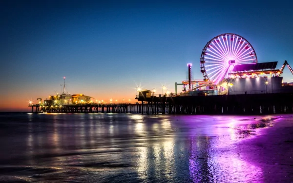 Night view of the illuminated ferris wheel and amusement park at Santa Monica Pier, Los Angeles, reflecting vibrant lights on the ocean shore.