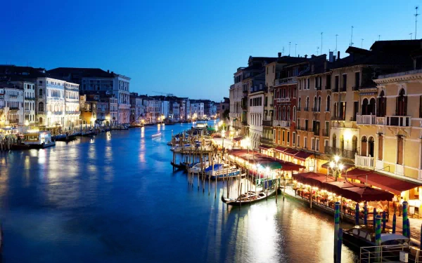 A stunning view of Venice at dusk, showcasing illuminated buildings along the canal, reflections in the water, and charming restaurants beside the waterways. A captivating Italian landscape.