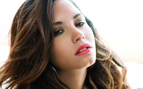 Close-up portrait of Demi Lovato with flowing hair and bold lips, set against a soft, light background. This image serves as an HD desktop wallpaper for music fans.