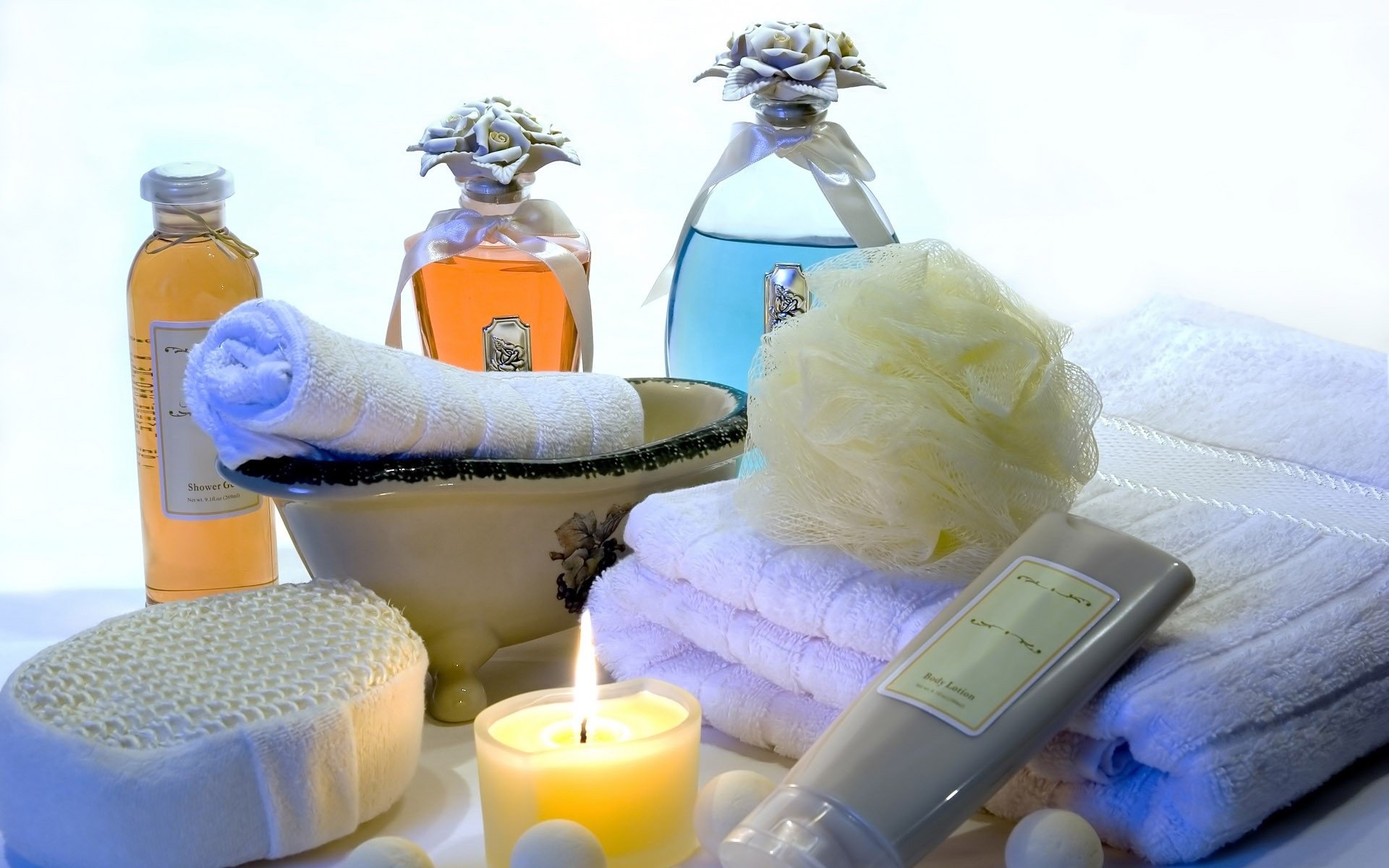 HD desktop wallpaper featuring a serene spa still life with a lit candle, rolled towel, bath pouf, lotion, and bottles arranged on a white surface.