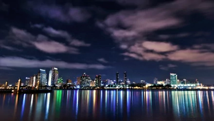 HD PC desktop wallpaper: man-made San Diego skyline at night, illuminated skyscrapers reflecting across the bay beneath sweeping clouds.