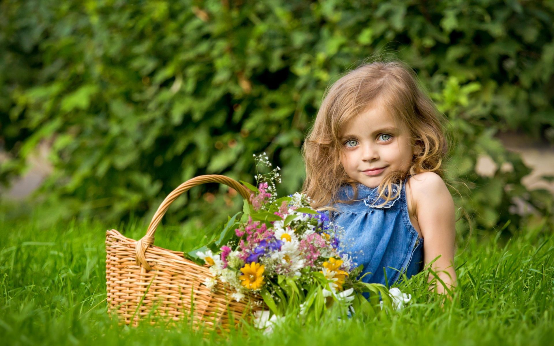 A young child with curly hair sits in lush green grass, smiling softly while holding a basket filled with colorful flowers, captured in vibrant HD photography for desktop wallpaper.