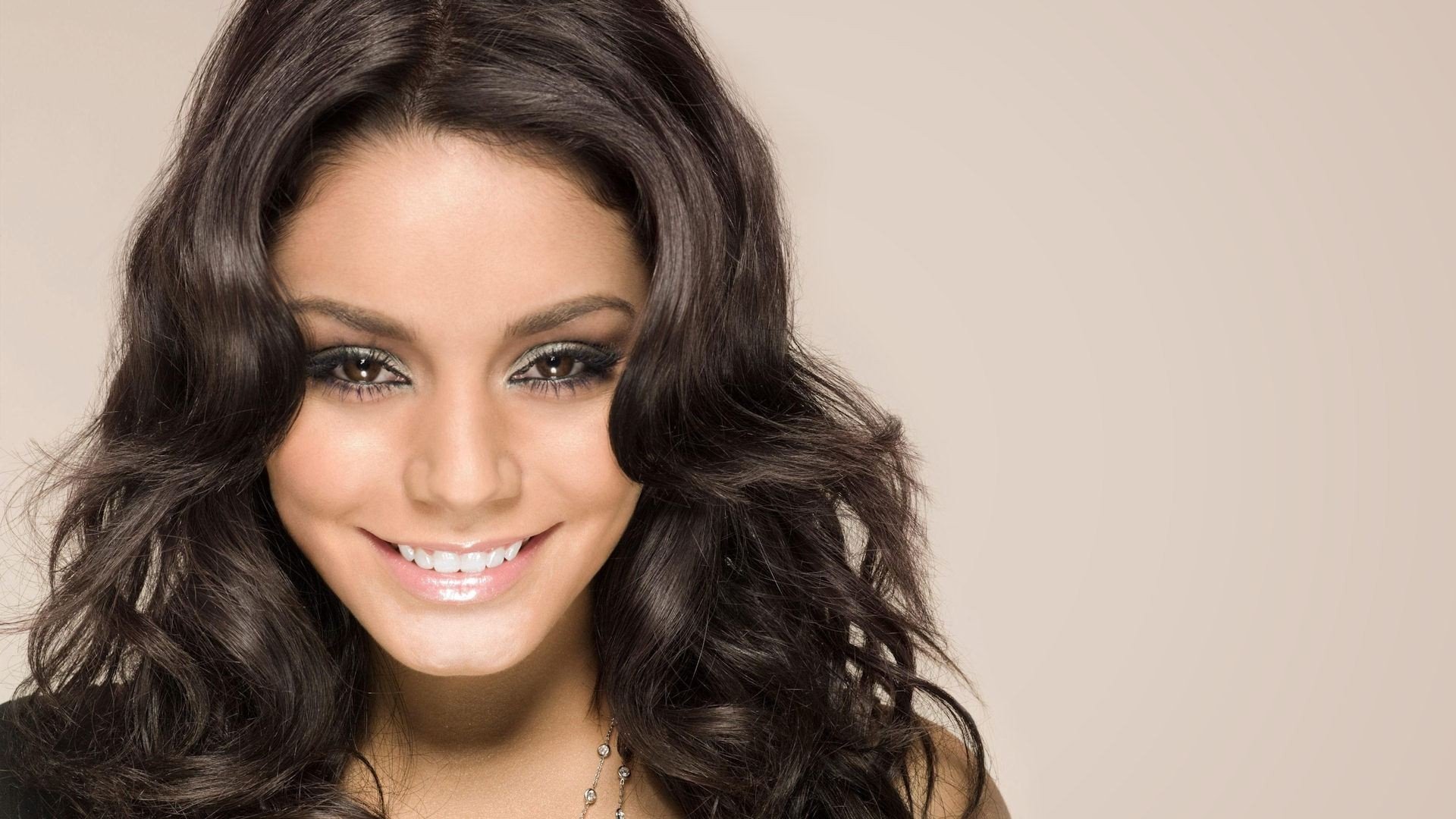 Close-up HD desktop wallpaper of celebrity Vanessa Hudgens smiling with wavy dark hair against a plain beige background.