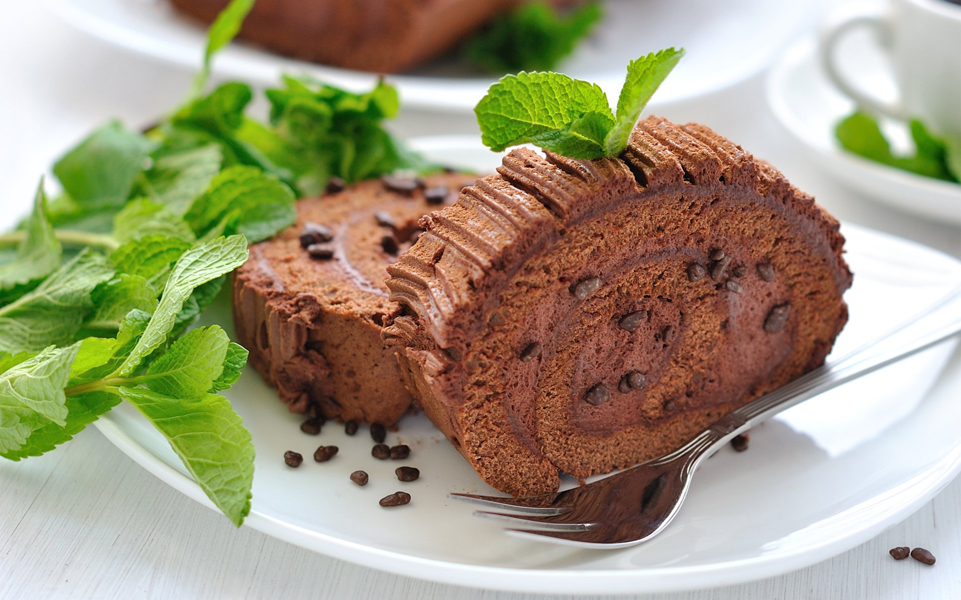 HD desktop wallpaper featuring a close-up of a chocolate rolled cake garnished with mint leaves and chocolate chips on a white plate.