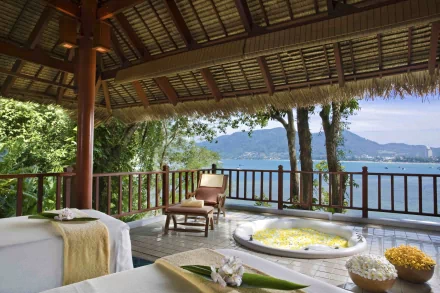 4K Ultra HD image of a man-made spa area with massage tables and a flower-filled tub, overlooking a serene waterside view framed by mountains and lush greenery.