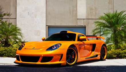 HD PC desktop wallpaper featuring a vibrant orange Porsche sports car parked outdoors against a modern building backdrop with palm trees.