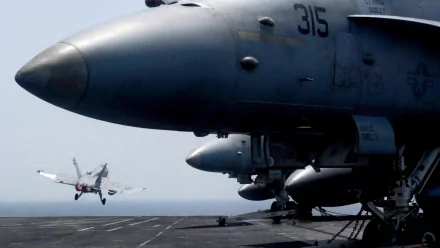 HD PC desktop wallpaper showing a military McDonnell Douglas F/A-18 Hornet nose on an aircraft carrier deck while another Hornet lands in the background.