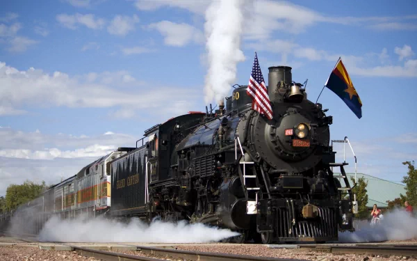 A classic steam train billows white smoke, adorned with American and state flags, set against a vibrant blue sky. This 4K Ultra HD image showcases a powerful vehicle in motion.