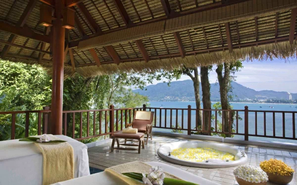 4K Ultra HD image of a man-made spa area with massage tables and a flower-filled tub, overlooking a serene waterside view framed by mountains and lush greenery.