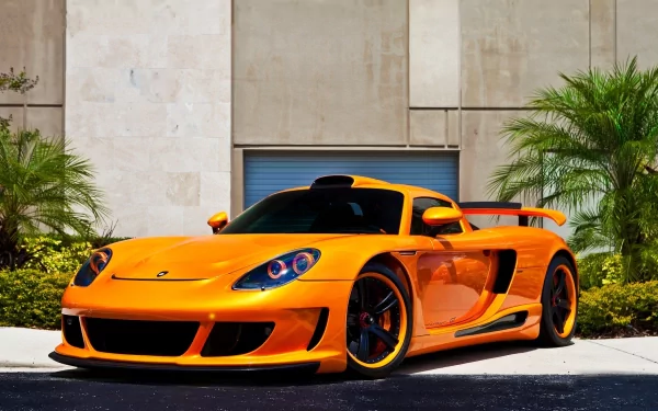 HD PC desktop wallpaper featuring a vibrant orange Porsche sports car parked outdoors against a modern building backdrop with palm trees.