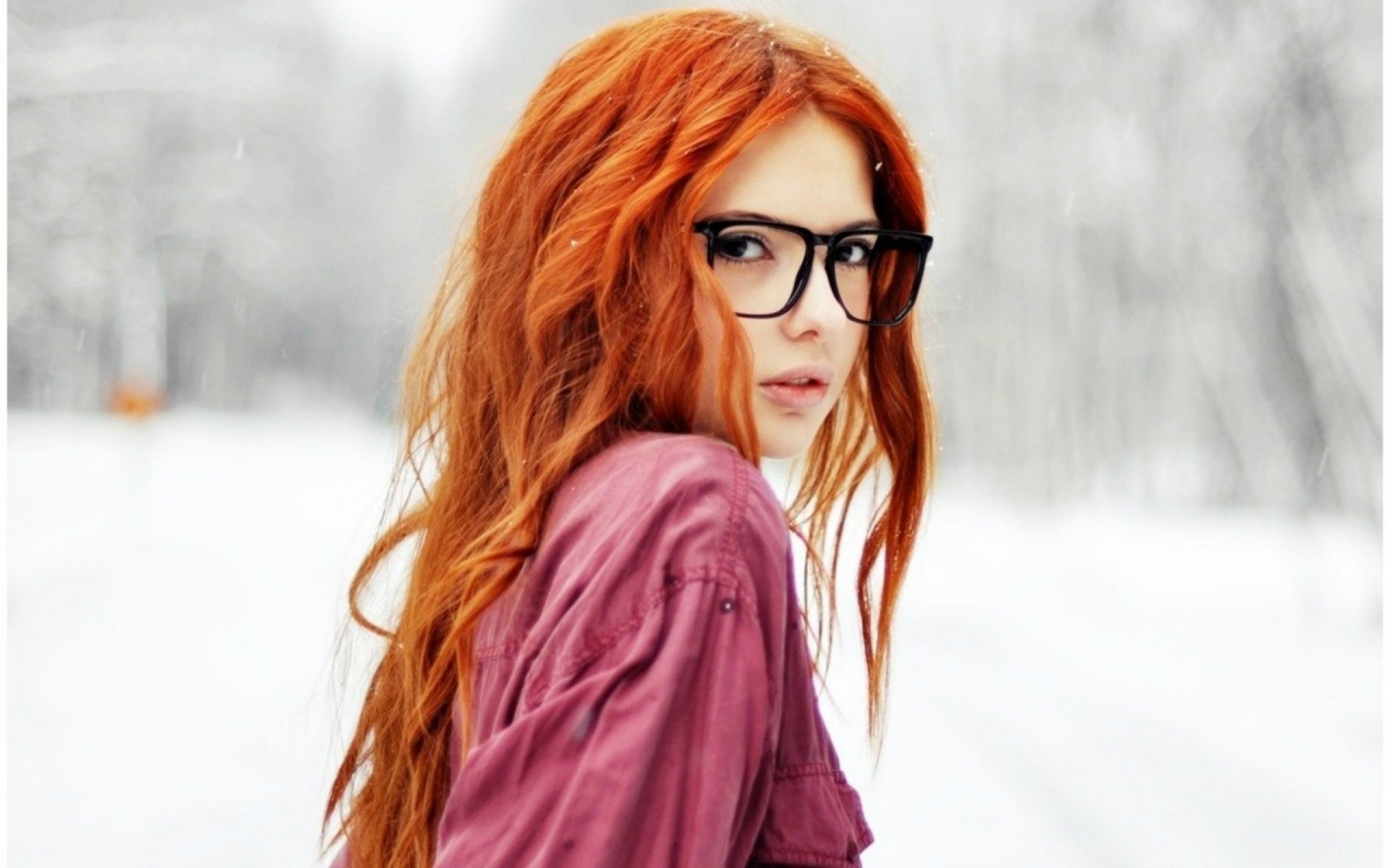 A beautiful redheaded woman with glasses gazes back over her shoulder, set against a snowy background. This HD desktop wallpaper captures her striking features and captivating eyes.