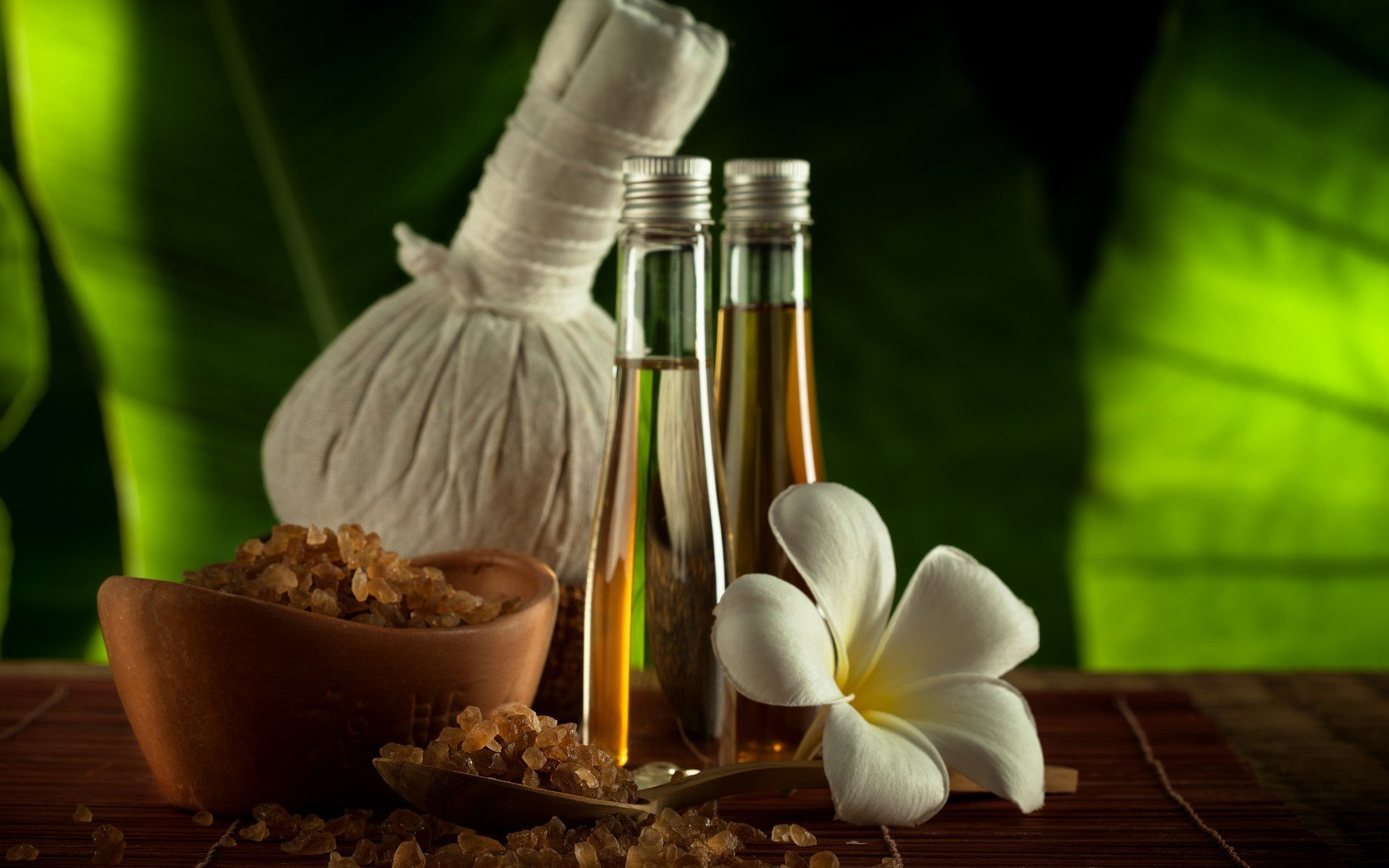 HD desktop wallpaper featuring a man-made spa setup with essential oils, herbal compress, bath salts, and a white frangipani flower against a green leafy background.