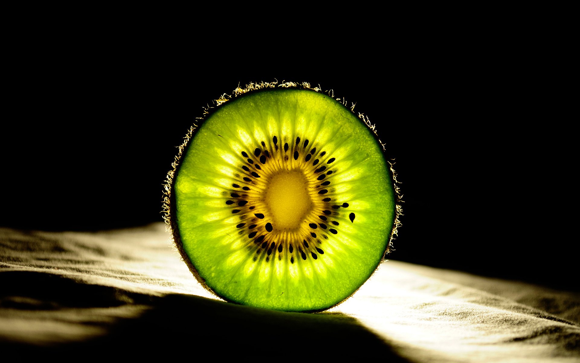 HD PC desktop wallpaper: close-up backlit sliced kiwi on a dark background, vibrant green flesh and radial seeds, a striking food image.