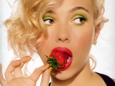 Scarlett Johansson playfully holds a strawberry to her lips, showcasing vibrant green eye makeup and bold red lips, creating a striking and colorful HD desktop wallpaper.