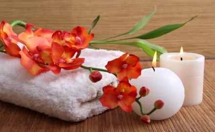 A serene spa setting featuring soft towels, vibrant orange orchids, and flickering candles, creating a soothing ambiance for relaxation. This HD wallpaper evokes tranquility and comfort.