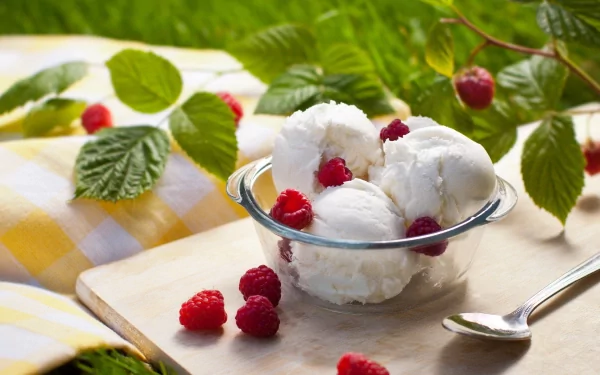 A delicious bowl of vanilla ice cream topped with fresh raspberries sits on a rustic wooden board, surrounded by green leaves and a checkered cloth, creating a vibrant dessert scene.