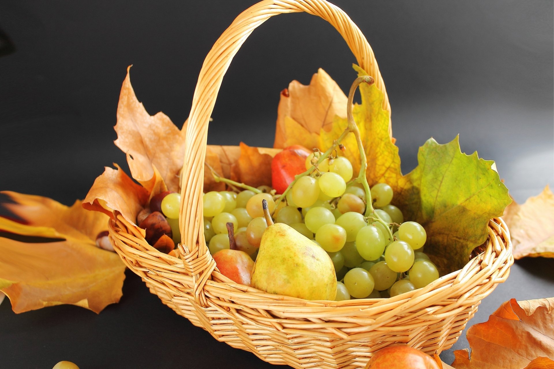 HD PC desktop wallpaper and background showing food: a wicker basket of fresh fruit (green grapes, pear) with autumn leaves on a dark backdrop.