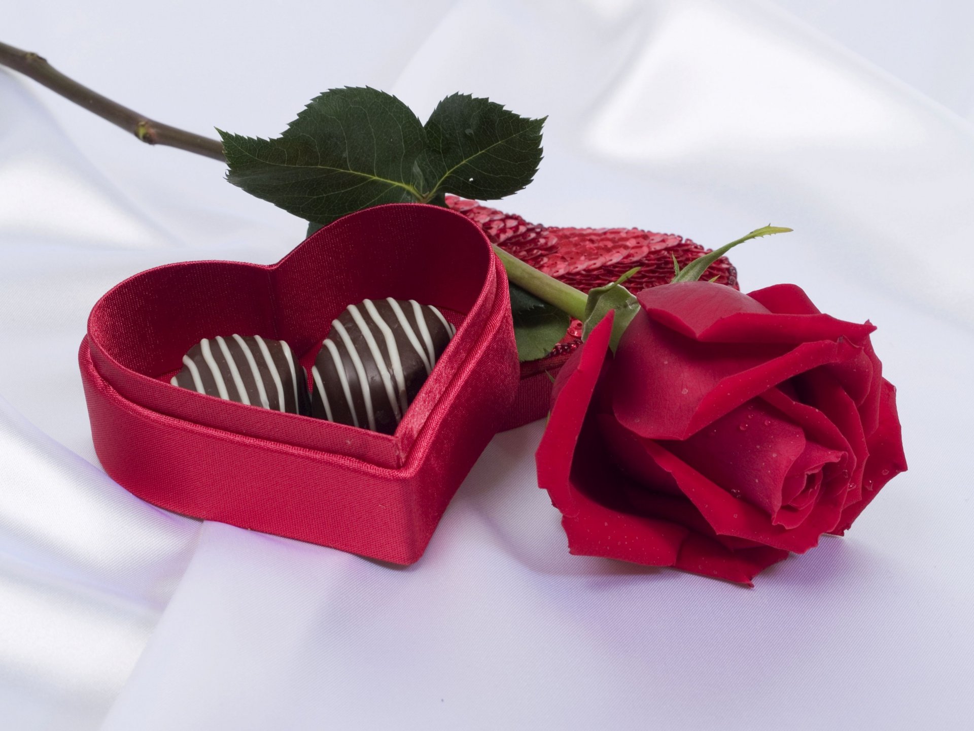A heart-shaped box of chocolate truffles beside a single red rose, set against a soft white background, capturing the essence of Valentine's Day. HD PC desktop wallpaper.