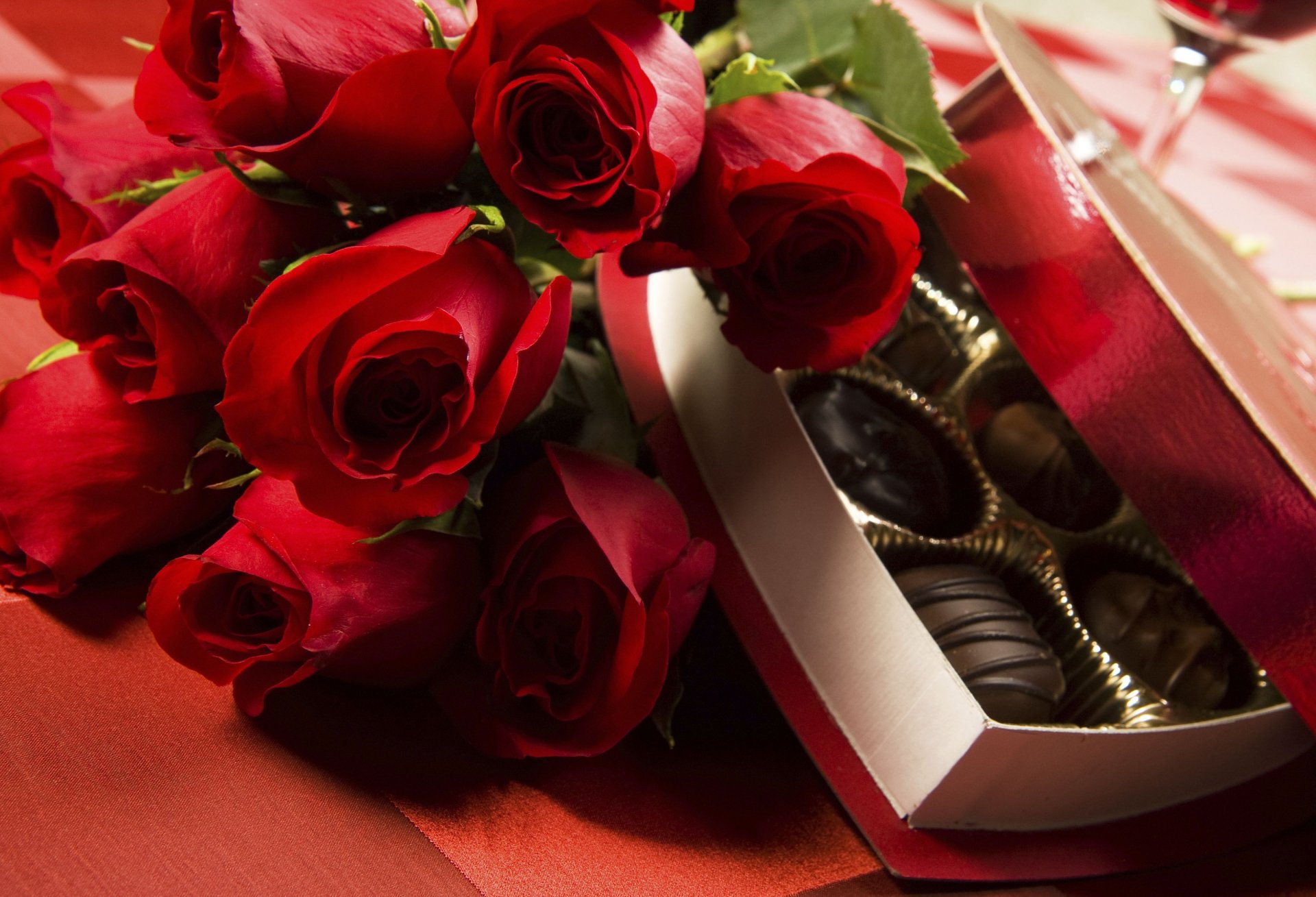 A romantic Valentine's Day scene features vibrant red roses beside a box of assorted chocolates, set against a warm, red background, creating an inviting holiday atmosphere.
