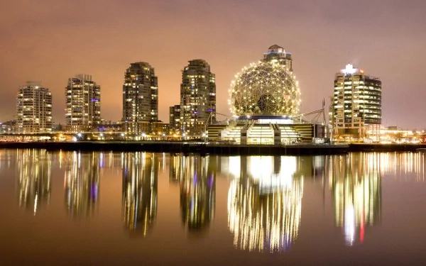 Nighttime cityscape of Vancouver, Canada, featuring illuminated modern buildings and the iconic geodesic dome reflecting on calm water.