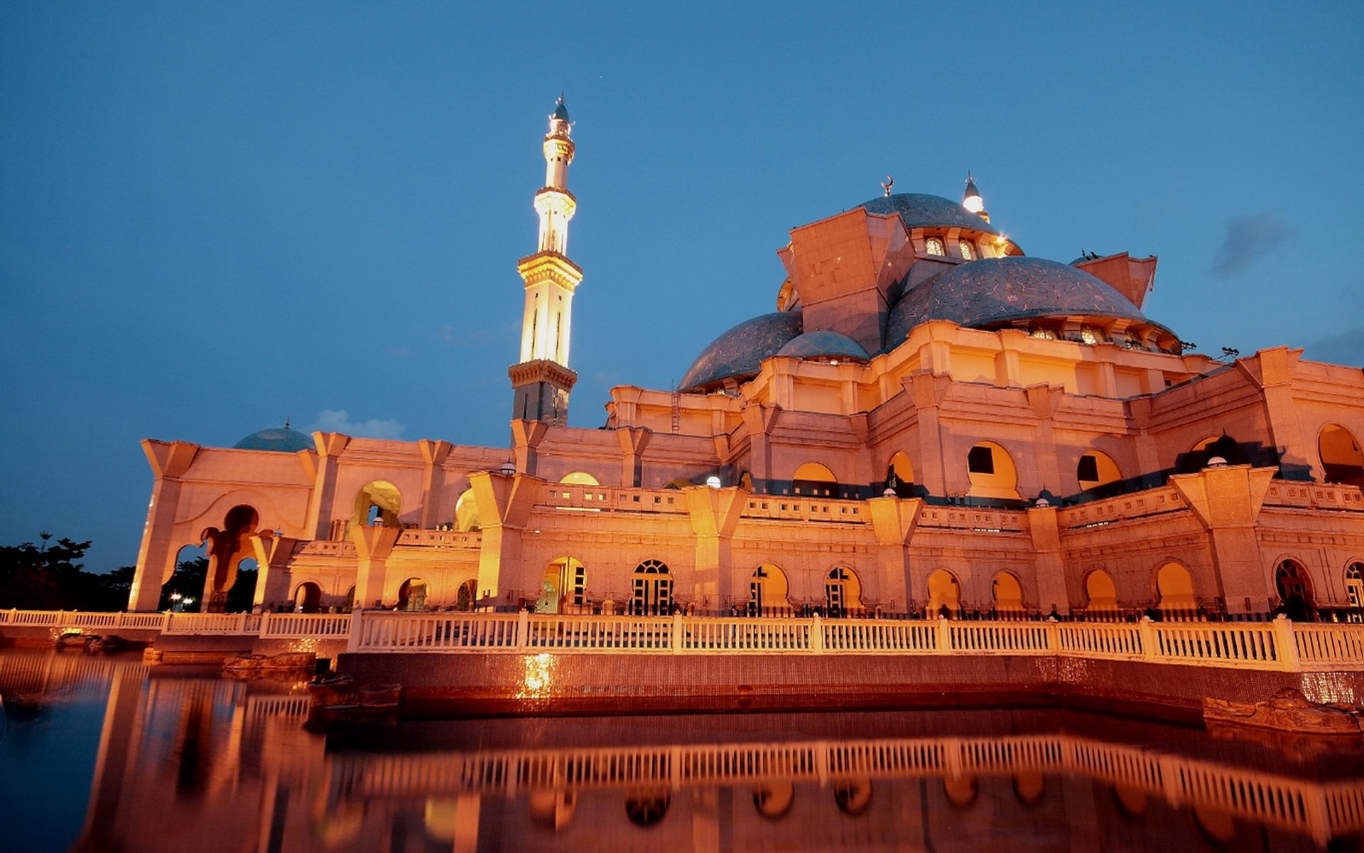 HD PC desktop wallpaper of the illuminated Federal Territory Mosque at dusk — ornate domes and minaret reflected in calm water, a religious landmark beneath a deep blue sky.