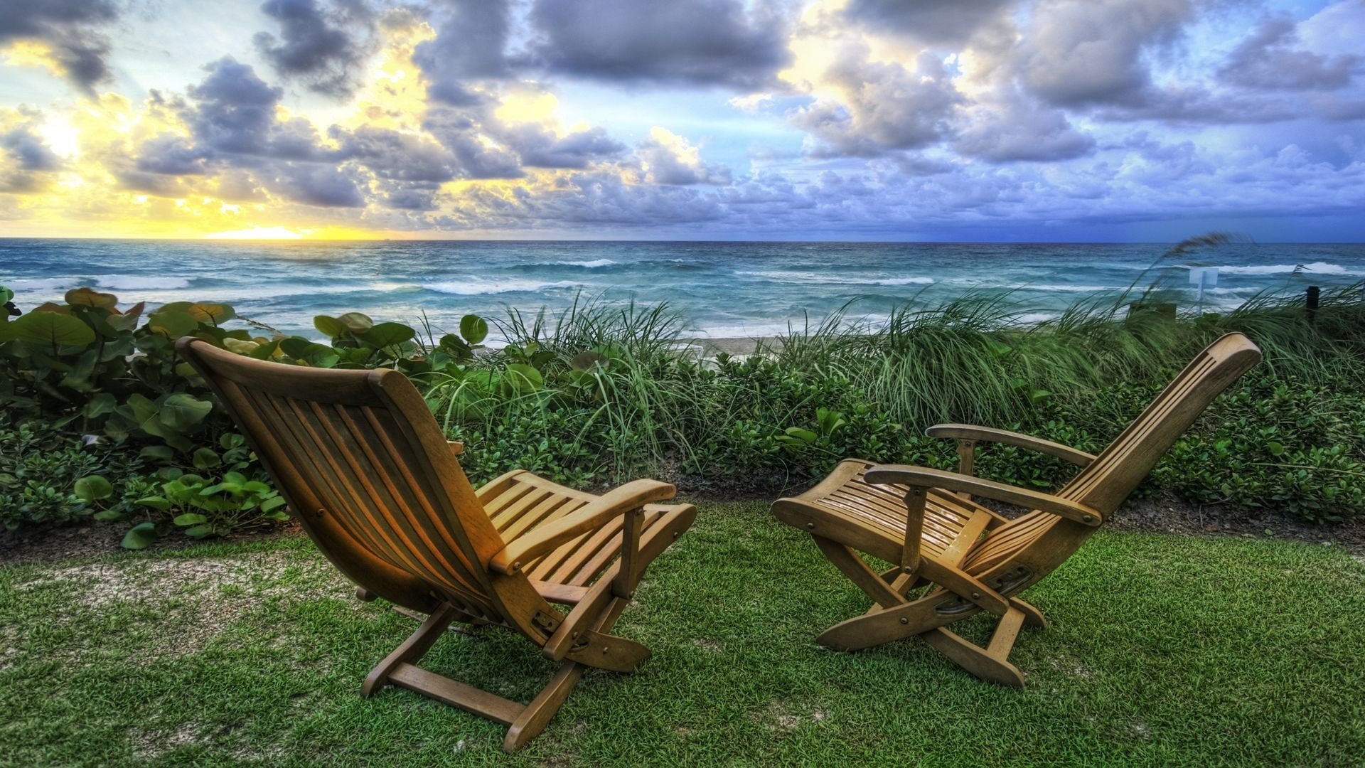 HD PC desktop wallpaper photography: two wooden chairs on a grassy dune facing a turquoise ocean under dramatic clouds, holiday seaside background.