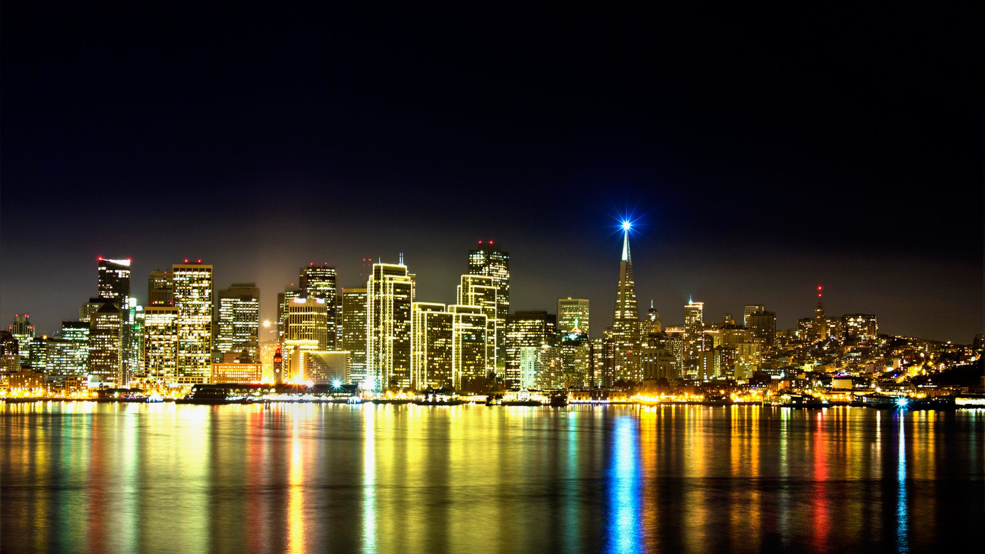 HD desktop wallpaper of San Francisco's illuminated skyline at night, showcasing its iconic man-made cityscape reflected on the water.