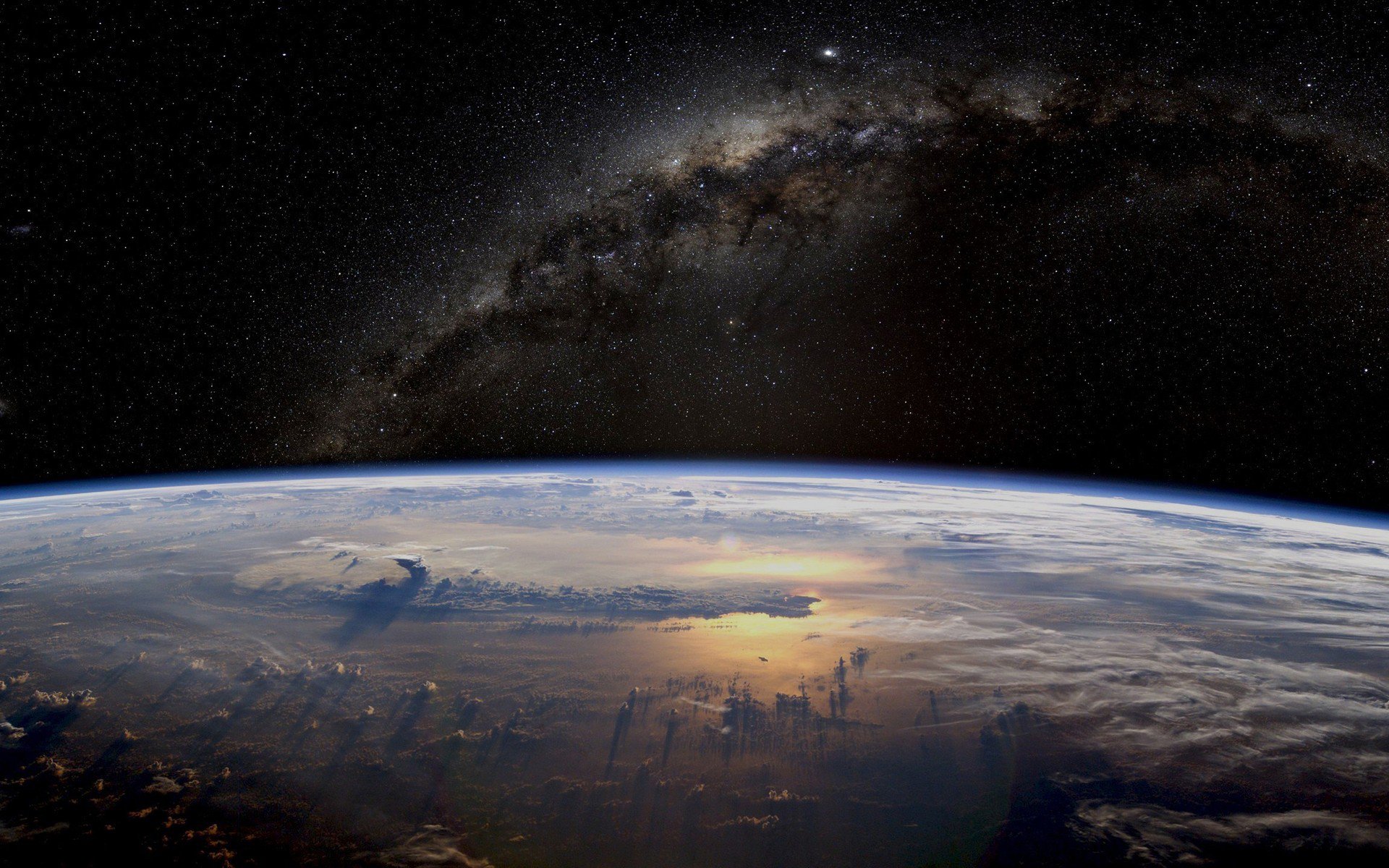Sci‑fi HD PC desktop wallpaper: curved planet horizon above a cloud layer, sunlit ocean reflections and shadowed cloud towers beneath a glowing Milky Way arching across a star-filled sky.