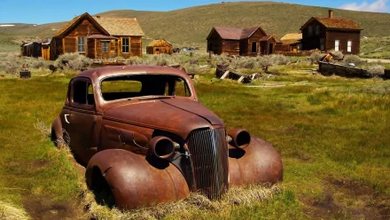HD PC desktop wallpaper showing a rusted vehicle wreck in a grassy field with old wooden houses and rolling hills under a clear blue sky.