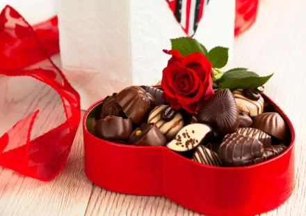 A heart-shaped box filled with assorted chocolates, topped with a red rose, sits beside a decorative white gift box, creating an inviting and romantic food-themed HD wallpaper.