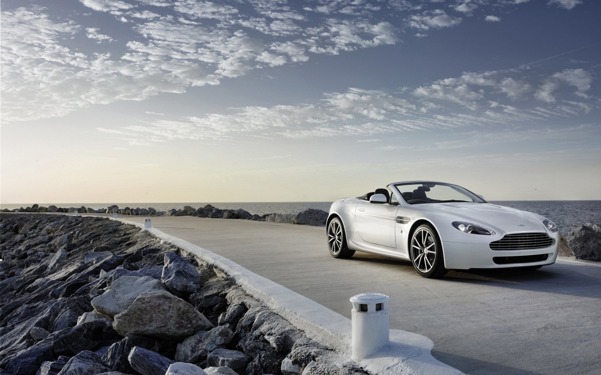 Silver Aston Martin Vantage convertible parked on a coastal jetty under a cloudy sky — 2K Quad HD PC desktop wallpaper/background.