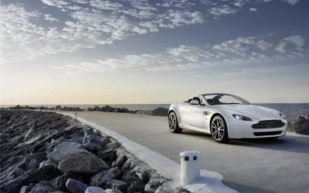 Silver Aston Martin Vantage convertible parked on a coastal jetty under a cloudy sky — 2K Quad HD PC desktop wallpaper/background.