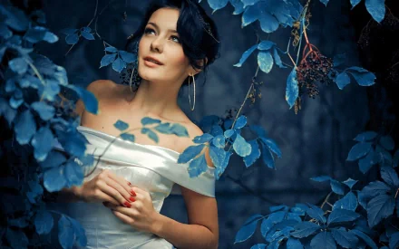 A bride dressed in an elegant white gown gazes thoughtfully amid lush blue foliage, capturing a moment of serene beauty in this HD desktop wallpaper.