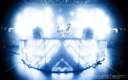 HD PC desktop wallpaper of a music DJ performing behind a glowing LED booth, bathed in intense blue-white stage lights and atmospheric fog.
