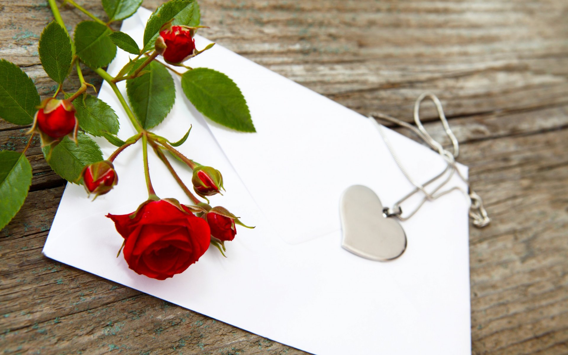 HD desktop wallpaper featuring a red rose, blank white envelope, and silver heart pendant on rustic wooden background, themed for Valentine's Day holiday.
