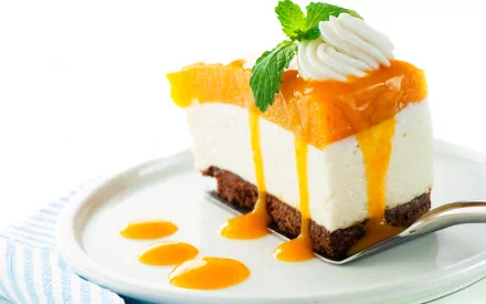 HD desktop wallpaper featuring a close-up of a slice of creamy cheesecake with an orange topping, drizzled sauce, and a mint garnish on a white plate.