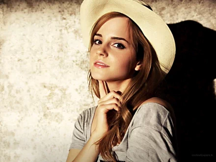 Emma Watson poses thoughtfully in this HD desktop wallpaper, wearing a light hat and a casual gray top against a textured background.