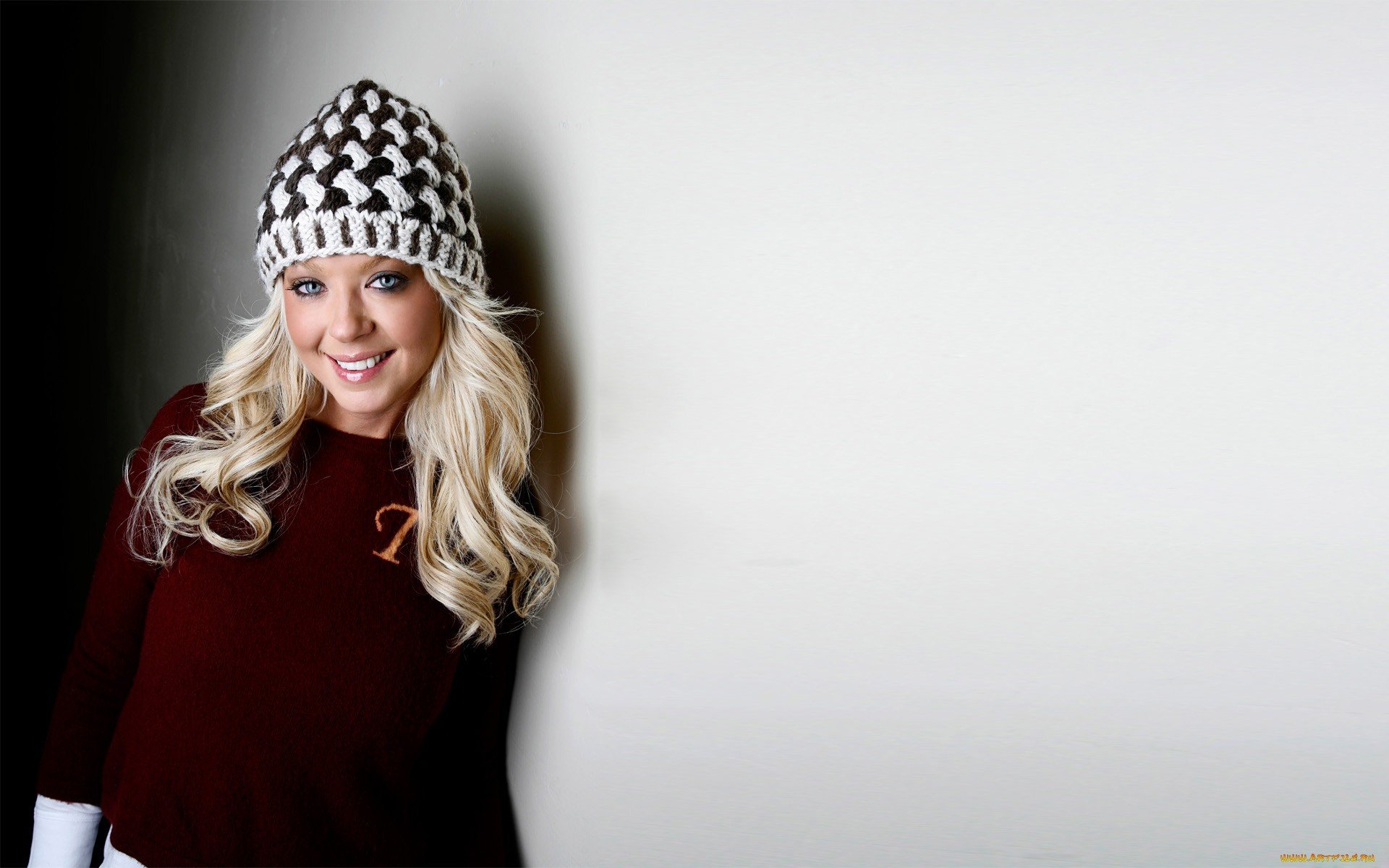 Tara Reid smiles warmly in this HD desktop wallpaper, wearing a stylish knit hat and cozy sweater, set against a minimalist background.