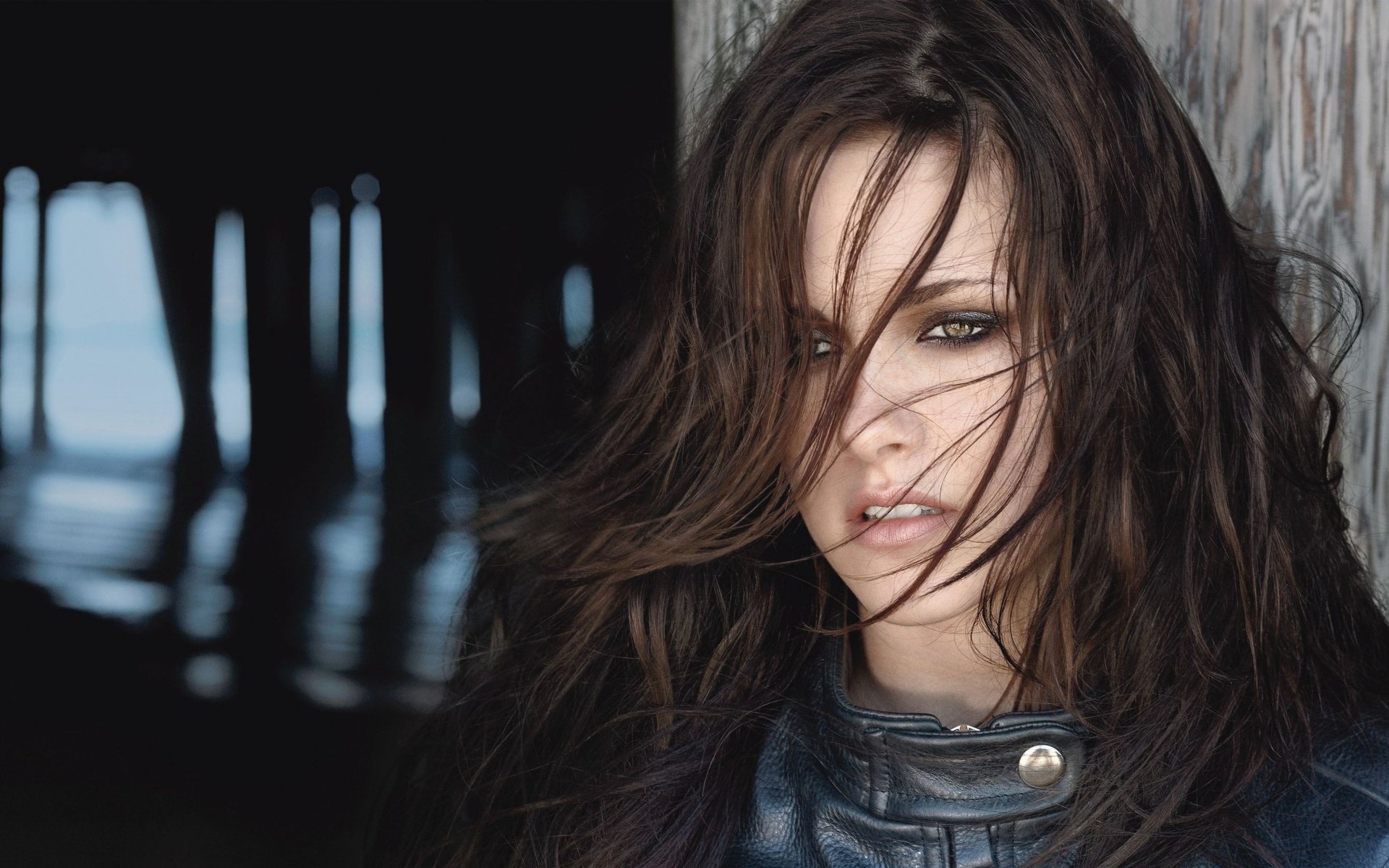 A striking HD wallpaper featuring celebrity Kristen Stewart with tousled hair and an intense gaze, set against a backdrop of wooden pillars and a blurred ocean view.