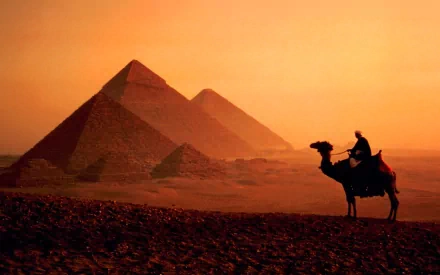 HD PC desktop wallpaper: silhouette of a camel rider before man-made pyramids at sunset, warm orange sky over desert dunes.