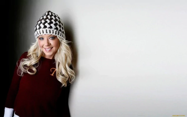 Tara Reid smiles warmly in this HD desktop wallpaper, wearing a stylish knit hat and cozy sweater, set against a minimalist background.