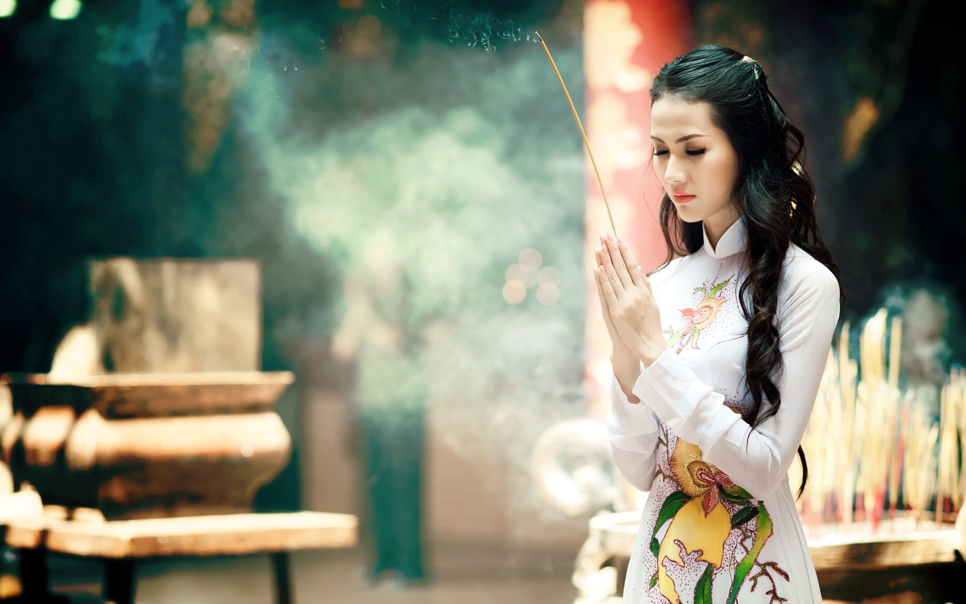 An Asian woman in traditional attire holds incense while praying, surrounded by a serene atmosphere filled with incense smoke. This HD wallpaper captures a moment of tranquility and reverence.