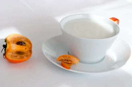 HD desktop wallpaper featuring a white cup filled with milk on a saucer, accompanied by a halved persimmon. The background is a clean, white surface, making the colors stand out. Tags: food, milk.