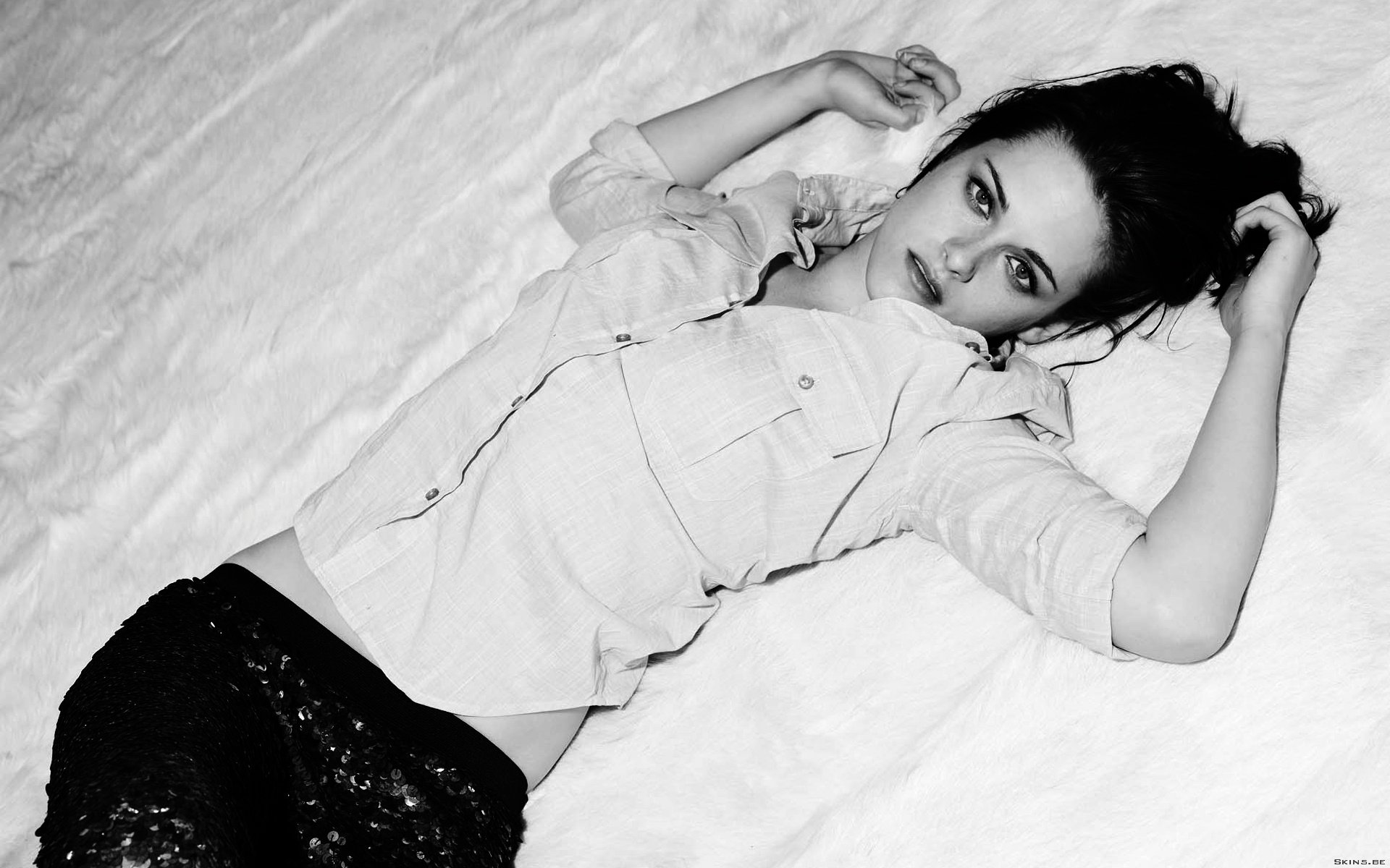 Black and white HD wallpaper of celebrity Kristen Stewart, lying on a textured surface, wearing a light shirt and dark pants, gazing thoughtfully at the camera.