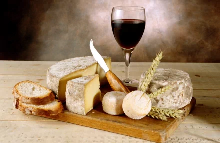 HD PC desktop wallpaper featuring a rustic cheese board with various cheeses, sliced bread, a knife, wheat stalks, and a glass of red wine on a wooden table.