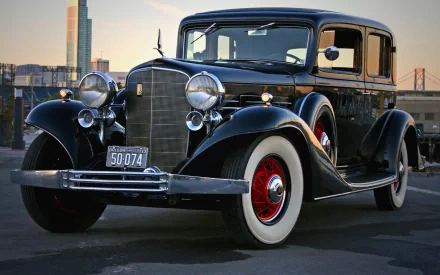 Black 1933 Cadillac V8 355C sedan vehicle with chrome grille and whitewall tires against a city dusk skyline, HD PC desktop wallpaper background.