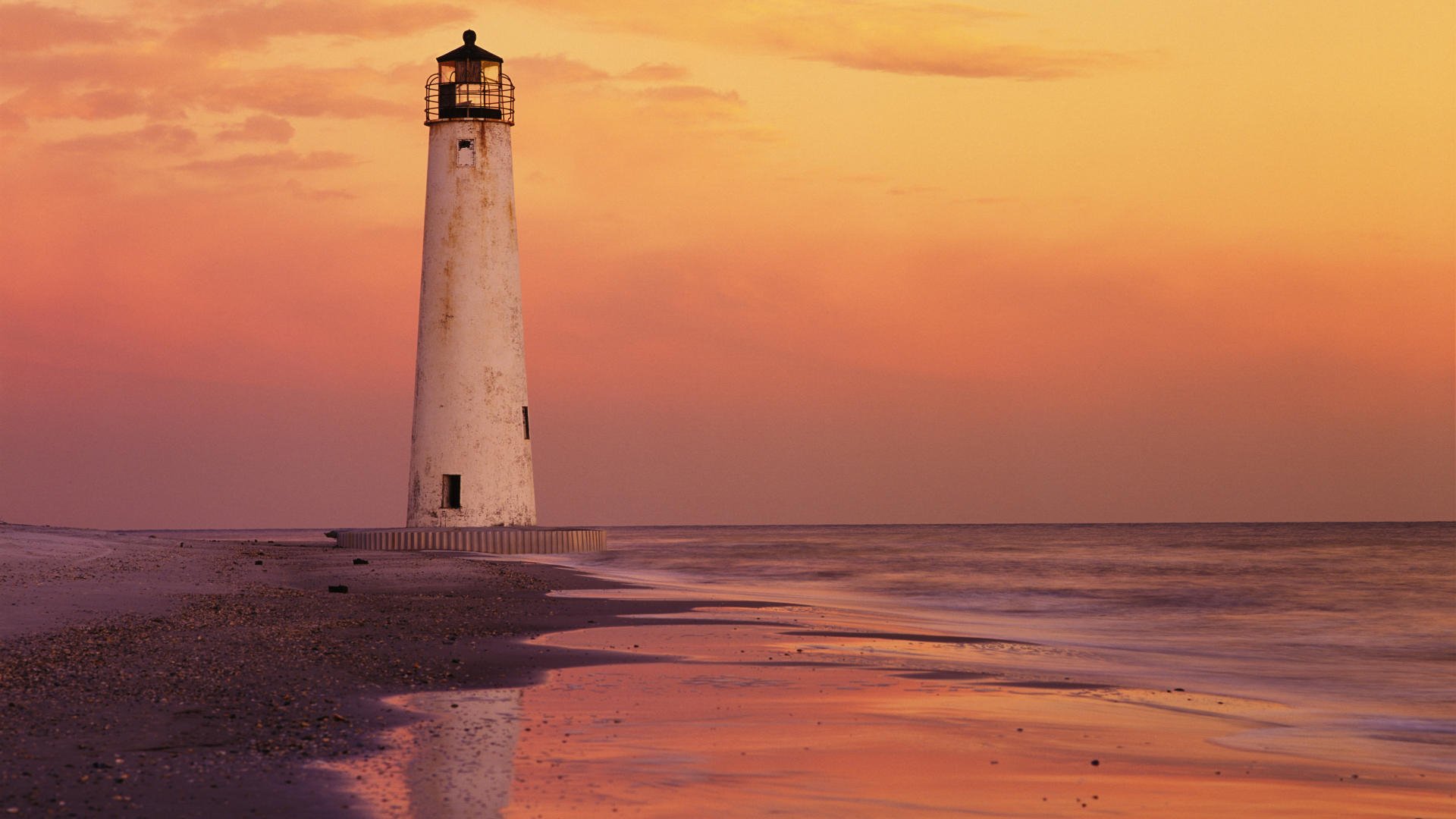 HD PC desktop wallpaper of a solitary man-made lighthouse on a sandy shore at sunset, warm orange sky reflected in wet sand.